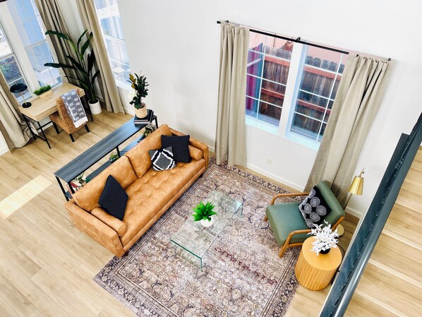 Sala de estar con sofá, una mesa de centro y una silla. Hay una ventana con cortinas, una planta y vistas a un edificio.