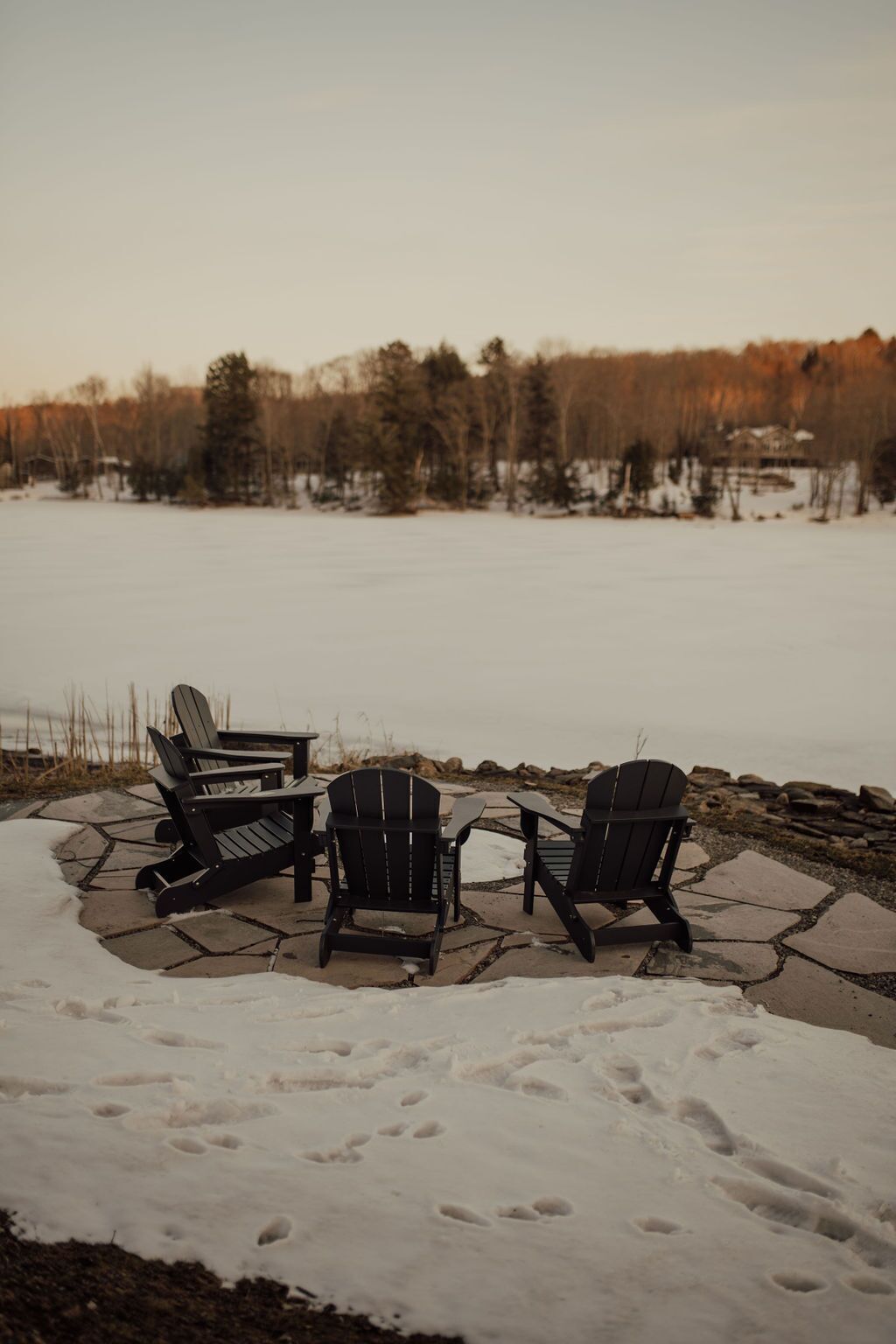A snow-covered patio with three Adirondack chairs and a table, overlooking a frozen lake with trees and a house in the background.