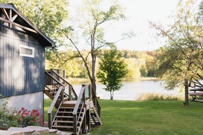 Eine Holzhütte mit einer Treppe, die zu einer Terrasse führt, mit Blick auf einen See, Bäume und einem Picknicktisch im Vordergrund.