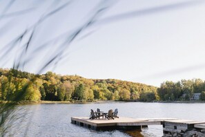 A wooden dock extending into a lake with chairs and a table, surrounded by trees and a house in the background.