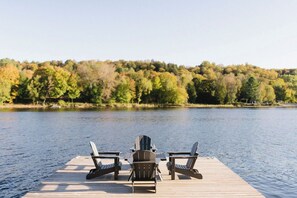Eine Holzstele mit zwei Adirondack-Stühlen, die auf einen ruhigen See ausgerichtet sind, umgeben von Bäumen mit Herbstlaub.