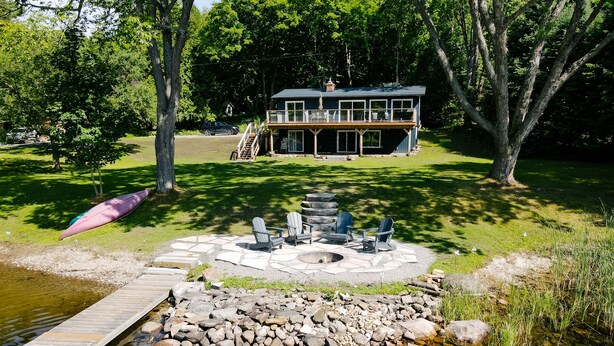 Eine Holzhütte am Seeufer mit einem Steinkamin und Adirondack-Stühlen.