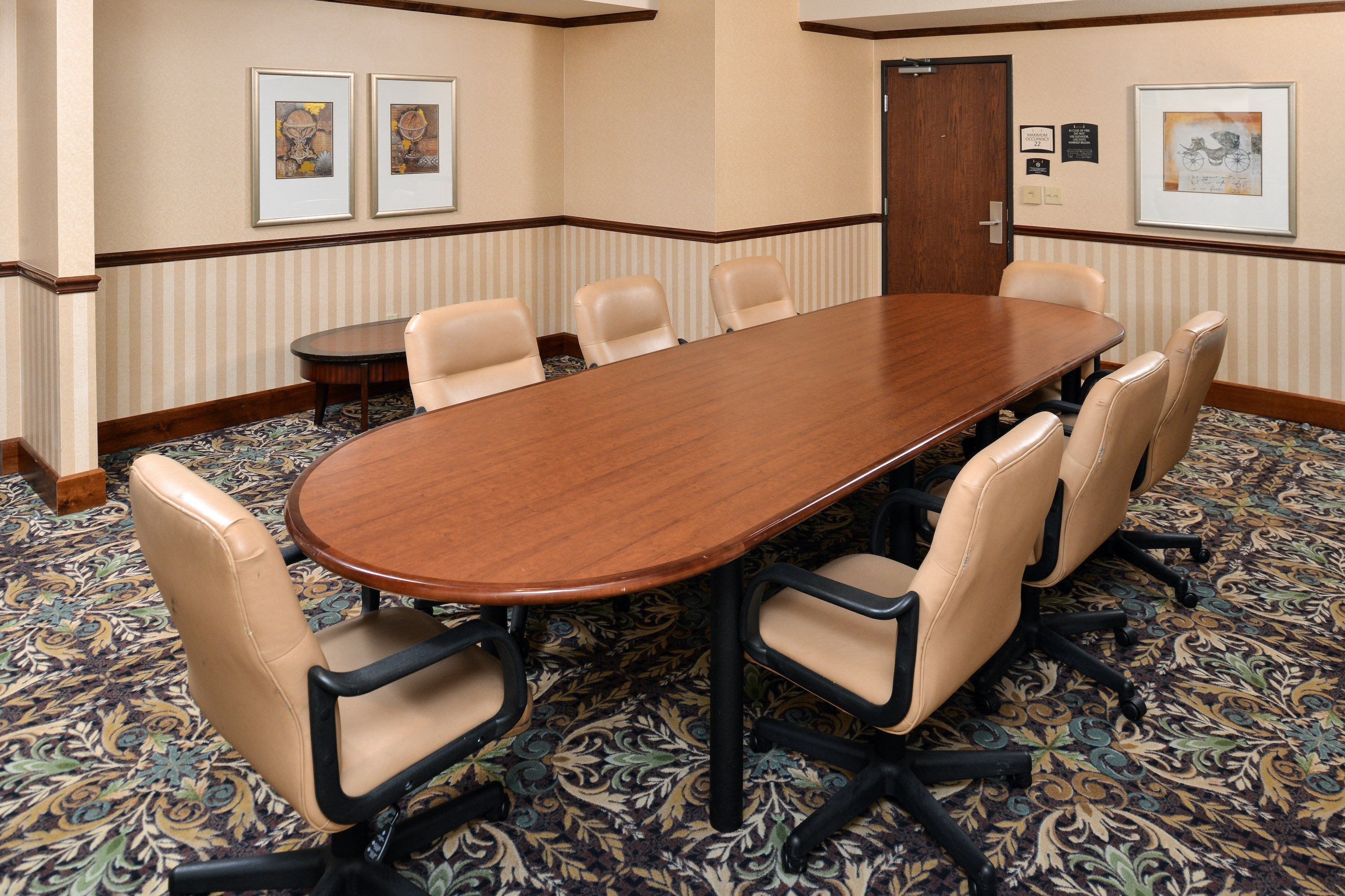 A conference room with a large wooden table surrounded by six beige chairs with black frames. The room has a patterned carpet, beige walls with a decorative border, and framed artwork on the walls.