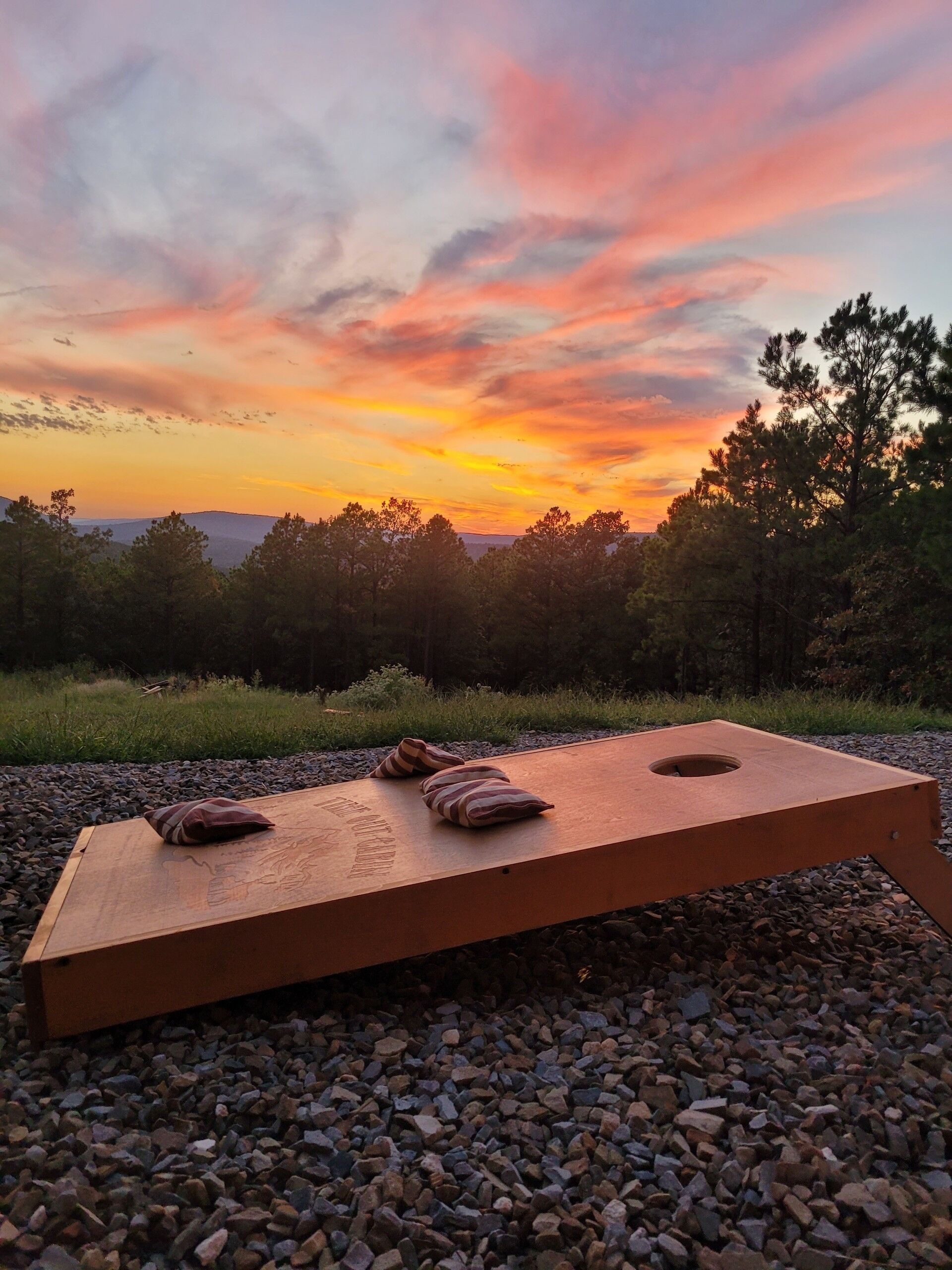 Un jeu de société en bois, installé sur une surface de gravier avec des coussins, entouré d’arbres et avec un coucher de soleil en arrière-plan.