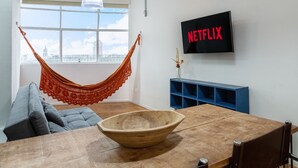 A living room with a hammock, a wooden dining table, a TV displaying "NETFLIX", and a view of a cityscape through a large window.