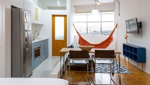 A modern kitchen with a stainless steel refrigerator, a dining area with a table and chairs, a hammock, and a TV mounted on the wall.