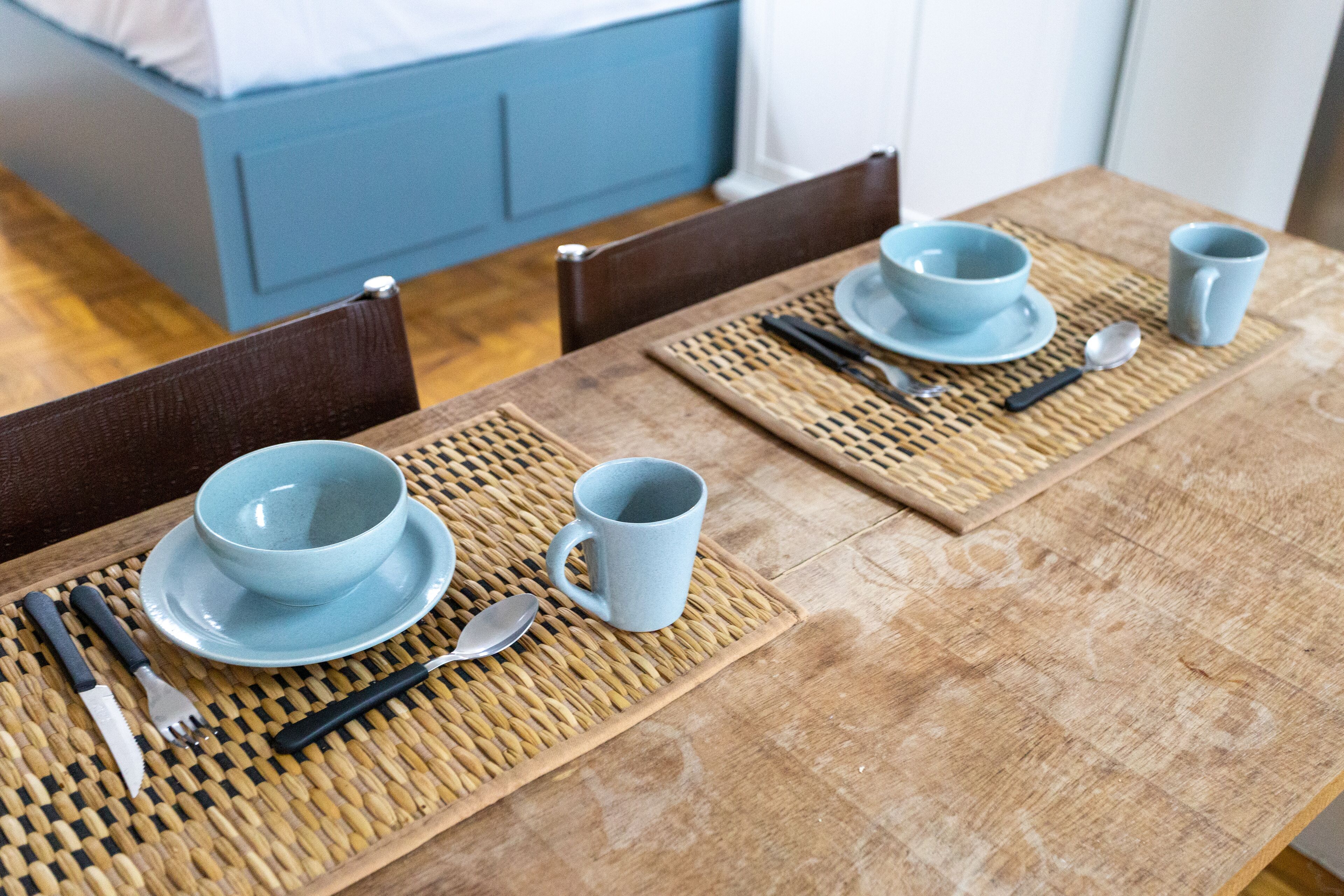 Een eettafel gedekt met blauwe borden, kommen en bekers, samen met bestek en een lepel, geplaatst op een geweven placemat.