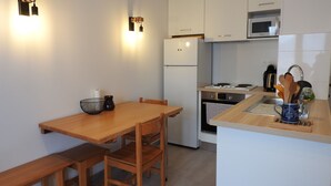 Una cocina compacta con gabinetes blancos, microondas, estufa y fregadero. Cuenta con una mesa de comedor de madera con sillas, y una pequeña encimera con cafetera y utensilios.