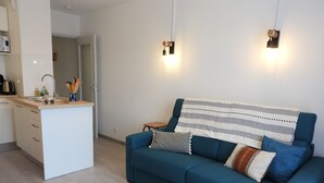 Una sala de estar moderna con un sofá azul, una zona de cocina con un lavamanos y armarios, y dos luces fijadas a la pared.