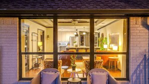 A cozy seating area with two chairs and a small table, a large window with a view of a kitchen and dining area, and a ceiling fan above. - Spacious Group Retreat | 3 Kings | Sleeps 25 (Ft. Worth)