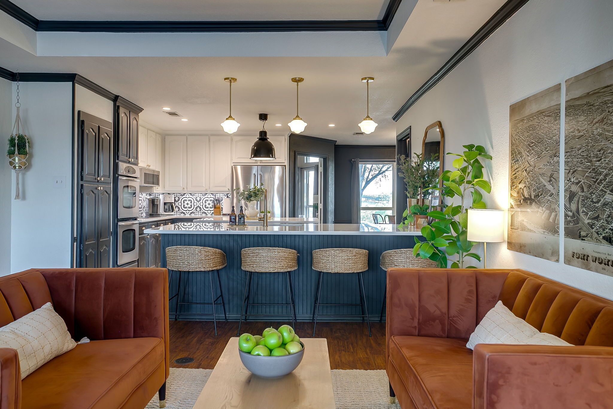 A modern kitchen with white cabinets, stainless steel appliances, and a central island. There are wicker bar stools, a brown leather sofa, and a bowl of green apples on a table.