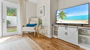 A living room with a large flat-screen TV on a white cabinet, a wicker chair with a blue cushion, a small side table with a plant, and a sliding glass door leading to a balcony with a view of palm trees and a beach.