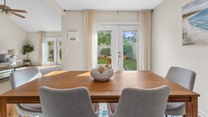 A dining area with a wooden table, grey upholstered chairs, a bowl of decorative items, and a view of the outdoors through large glass doors.