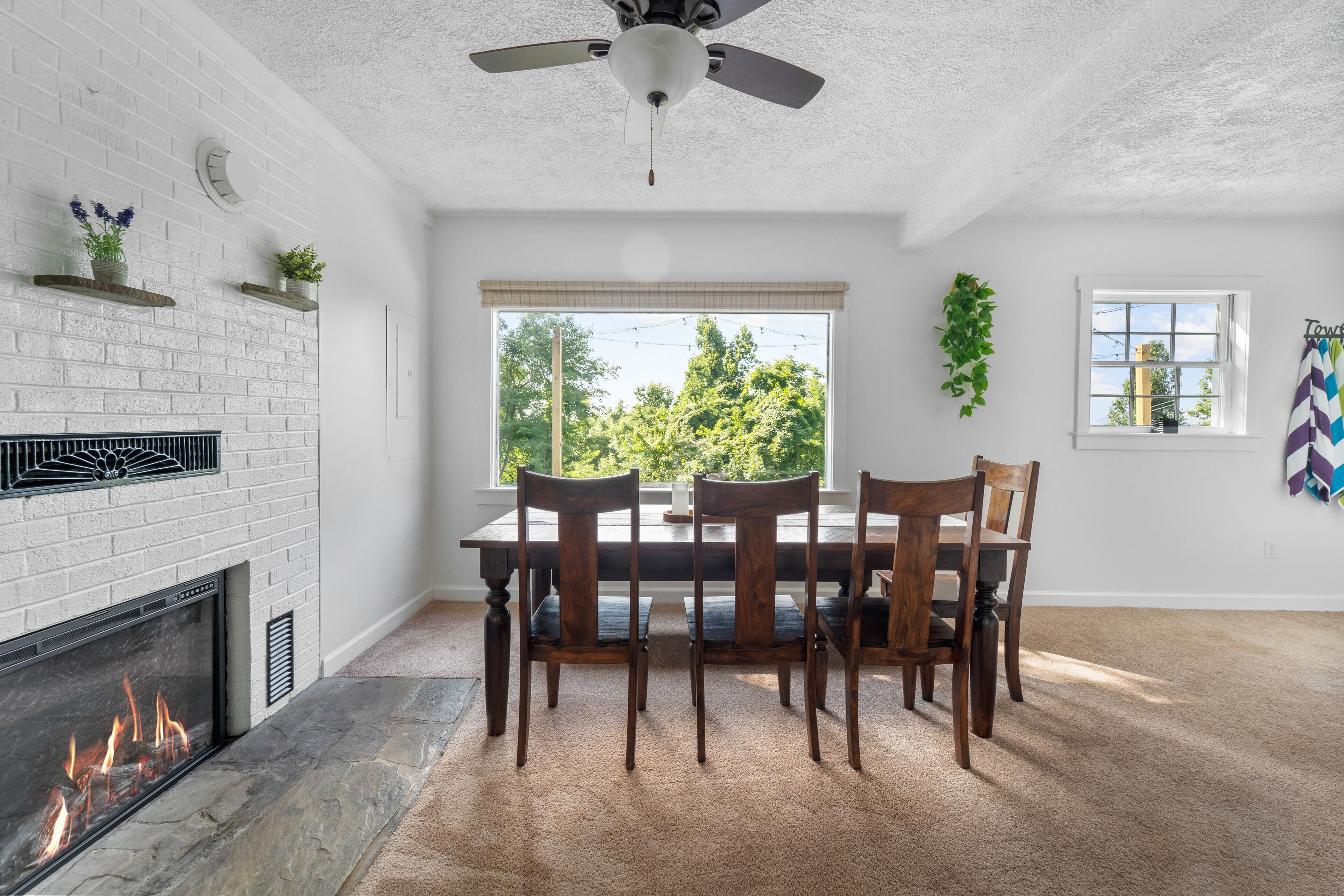 A dining area with a brick fireplace, a ceiling fan, and a view of trees through a large window.