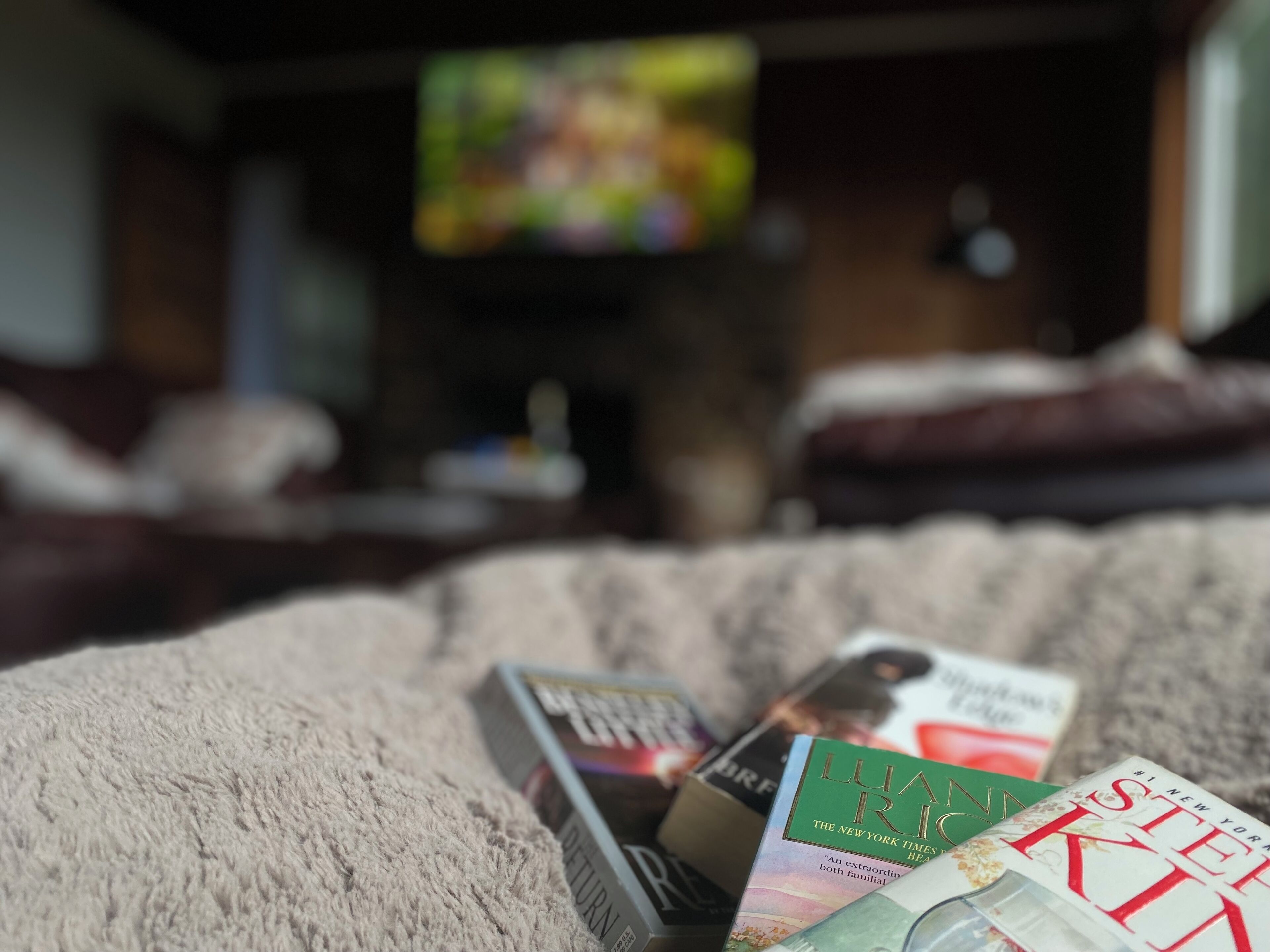 A collection of books on a textured surface, likely a couch or bed, with a television in the background.