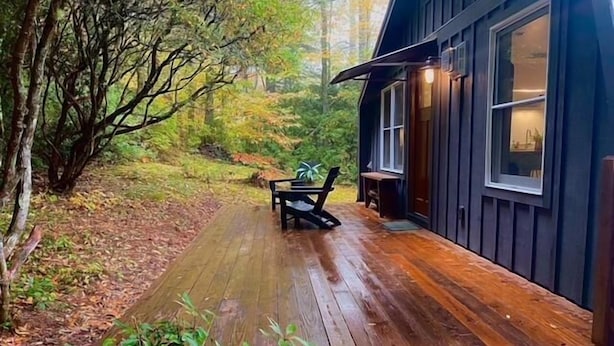 Une terrasse en bois menant à un chalet équipé d’un banc et d’une table, le tout entouré d’arbres.