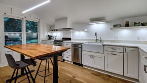 Une cuisine moderne avec des meubles blancs, une table à manger en bois et une vue sur les arbres à travers de grandes fenêtres.
