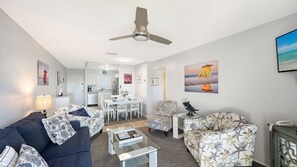 A living room with a sofa, armchair, coffee table, and a ceiling fan. There is a dining area with a table and chairs, and a kitchen in the background. - Condo# 205 Siesta Key Gulf view,steps to sand (Sarasota)