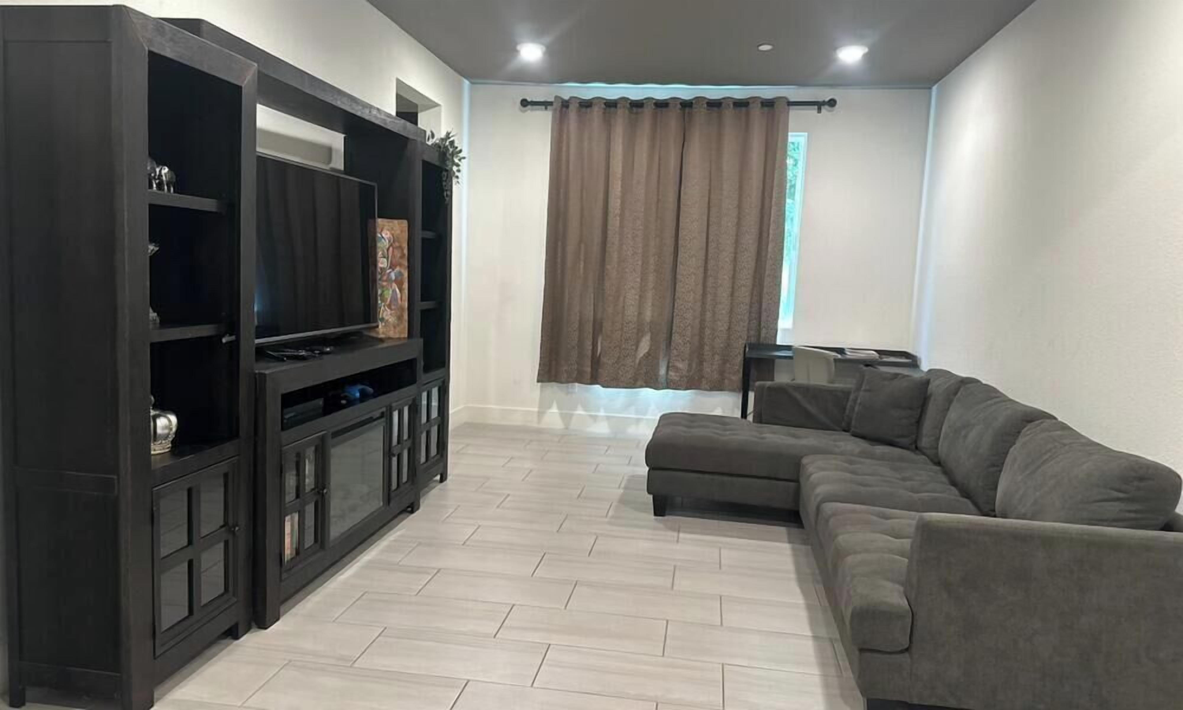 A modern living room with a dark wooden entertainment unit, a grey sectional sofa, and a large window with brown curtains.