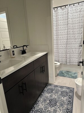 Een badkamer met een wit wastafelmeubel, donkere kastjes, een kamerkleed met dessin en een douchegordijn.