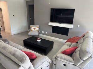 Een woonkamer met een aan de muur bevestigde televisie, een zwarte salontafel, twee banken met rode kussens, en een fauteuil met een patroon.