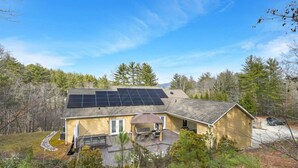 Una casa de dos pisos con paneles solares en el techo, un porche cubierto y un paisaje boscoso de fondo.
