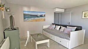 Ein modernes Wohnzimmer mit einem weißen Sofa, einem kleinen Tisch, einem Fernseher und einem Gemälde mit Strandmotiv.