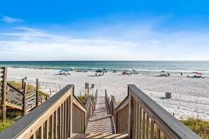 Un escalier en bois mène à une plage de sable où se trouvent des parasols et des gens.
