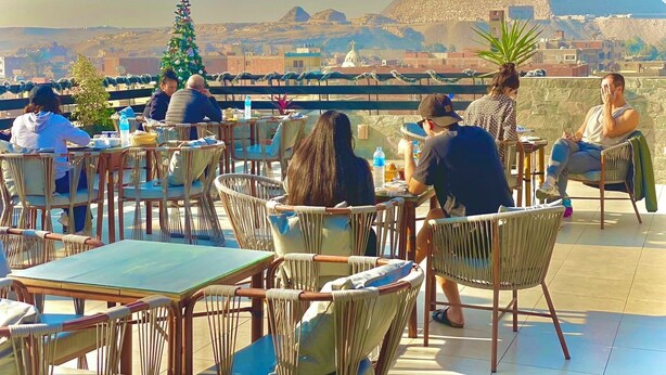 A rooftop dining area with wicker chairs and tables, overlooking a pyramid and cityscape. - Kemet Boutique Hotel (Giza)