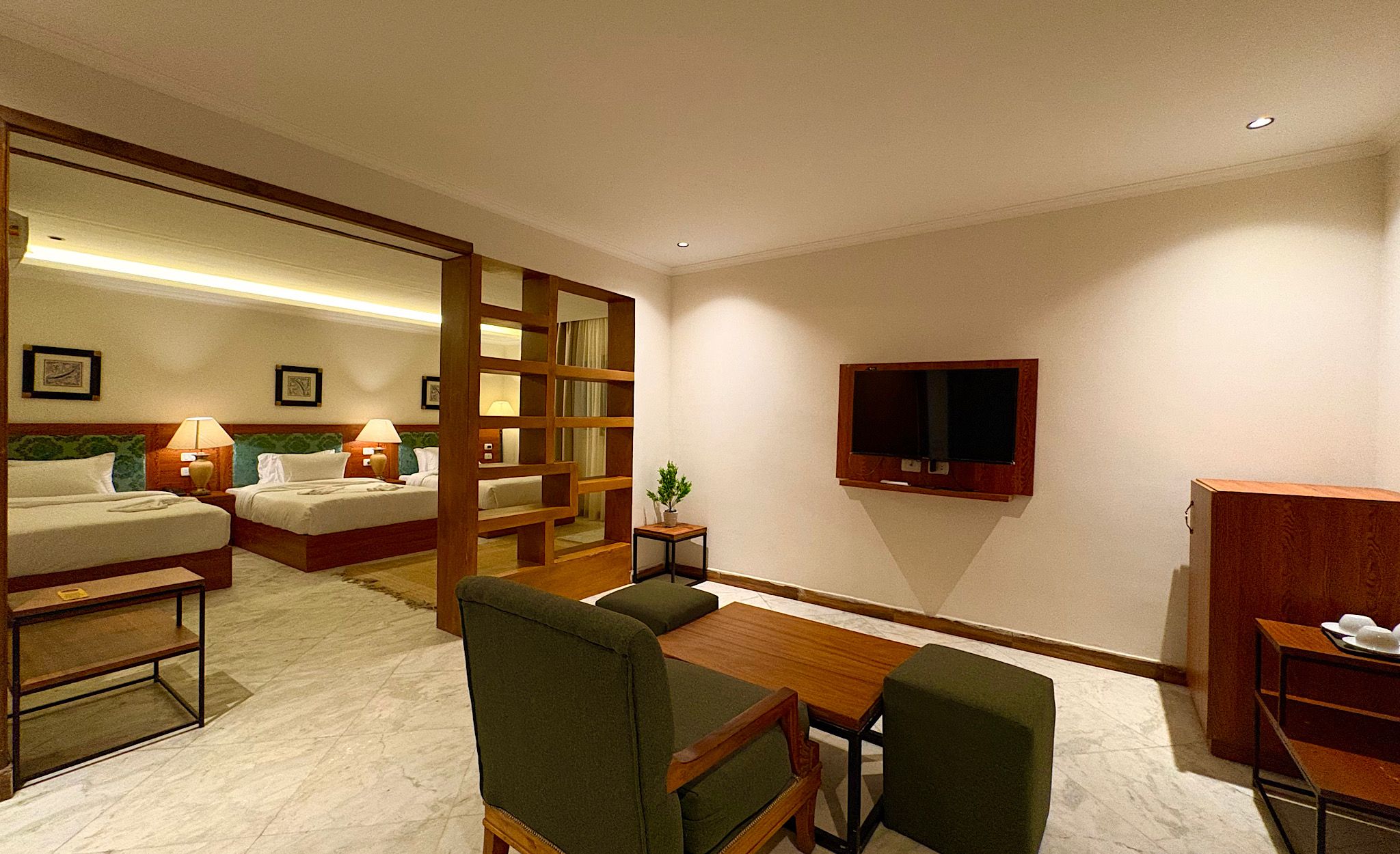 Habitación de hotel con cama, dos mesitas de noche con lámparas, televisor montado en la pared, una pequeña mesa con sillas y un armario de madera.