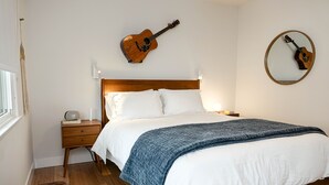 Ein Schlafzimmer mit einem hölzernen Bett, weißer Bettwäsche, einer blauen Decke, einer Gitarre an der Wand, einem Nachttisch mit einer Lampe und einem Beistelltisch.
