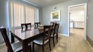 Comedor con mesa y sillas de madera, ventana con persianas, lámpara de techo y una zona de cocina con armarios.
