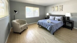 Un dormitorio con una cama, una silla, una mesita de noche, una lámpara y una ventana.