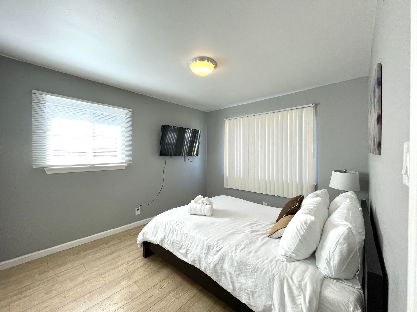 A bedroom with a bed, a TV mounted on the wall, and two windows with blinds.
