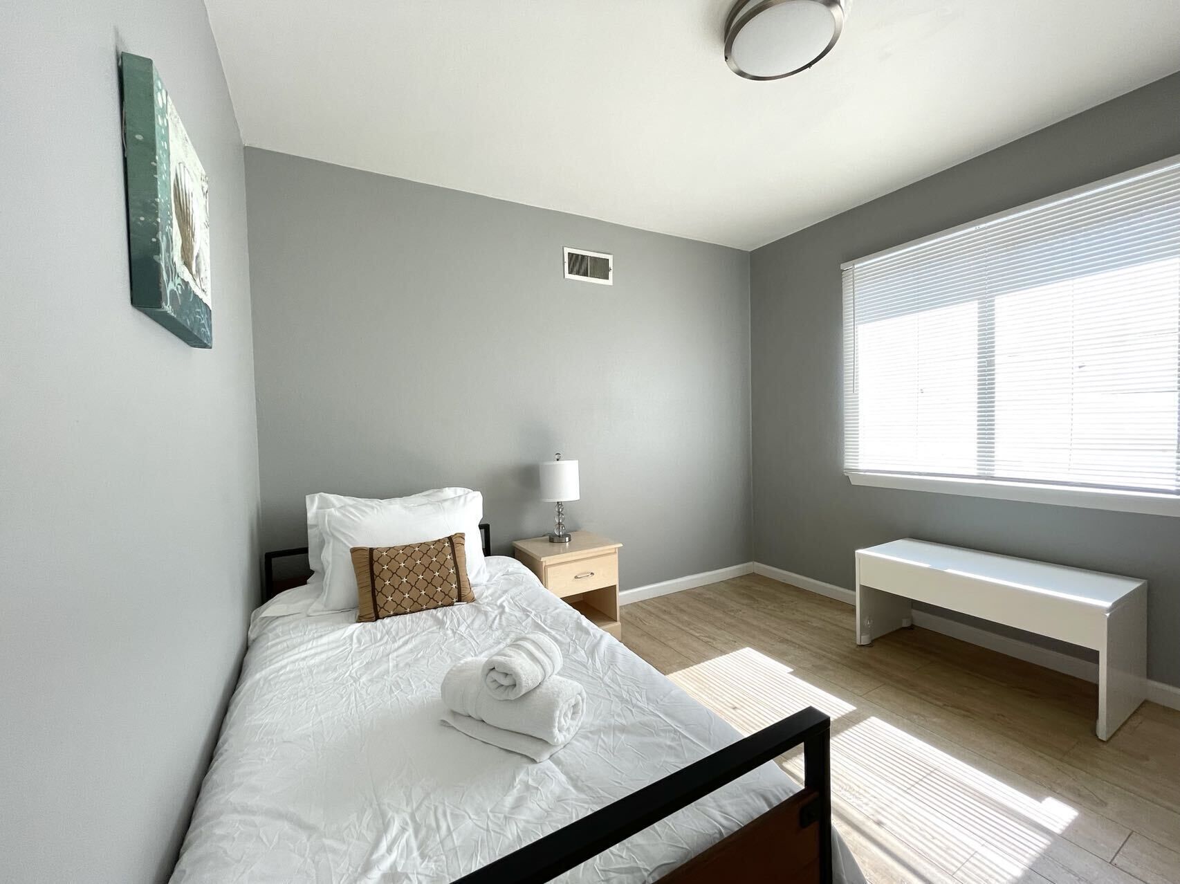 A bedroom with a bed, bedside table, lamp, bench, and a window with blinds.