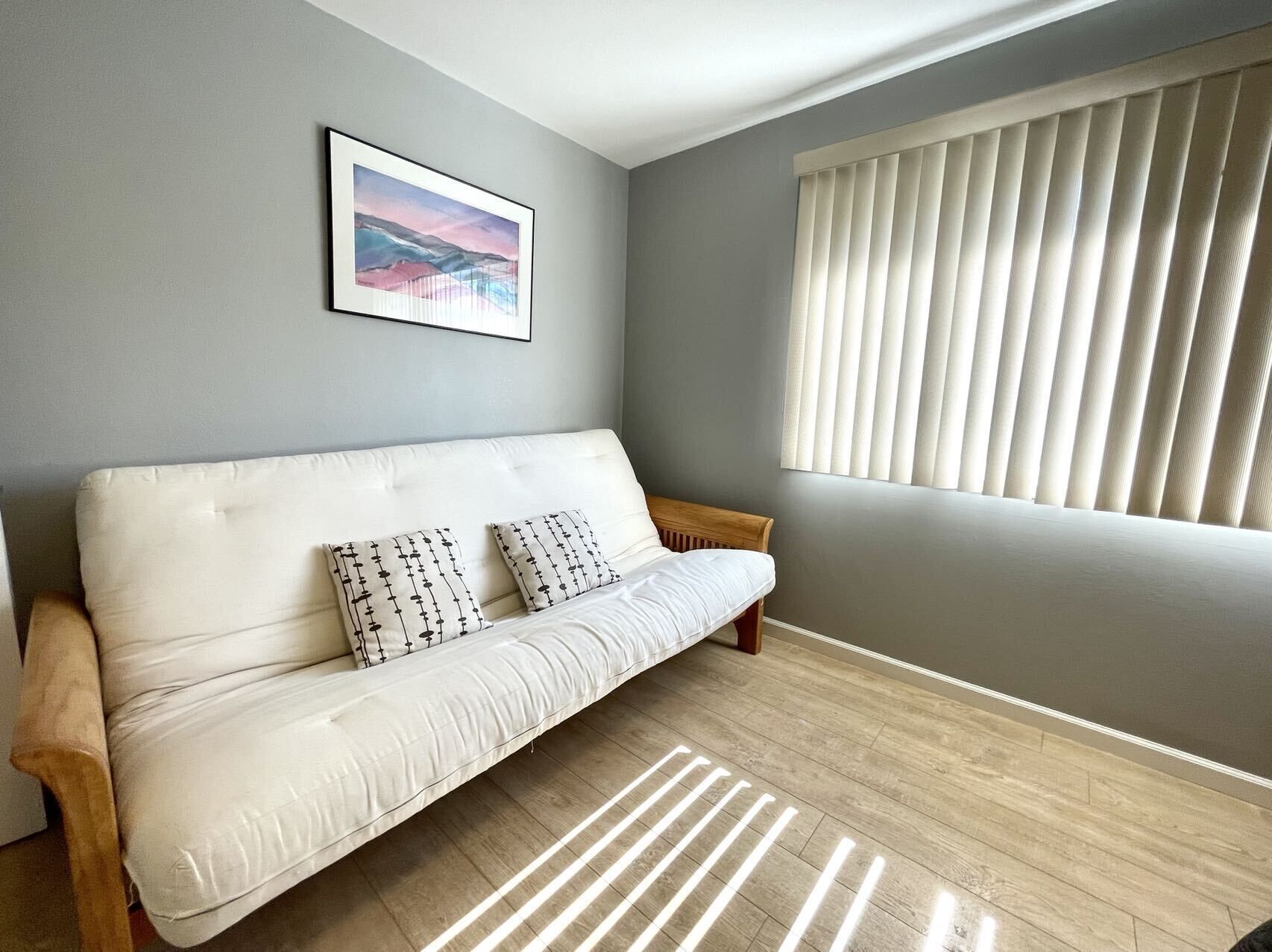 A modern living room with a white sofa, two patterned pillows, a framed picture on the wall, and a window with blinds.