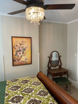 Quarto com cama, ventilador de teto, lustre, pintura floral emoldurada, cômoda de madeira e espelho.
