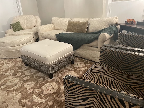 Uma sala de estar com um tapete estampado, um sofá branco com almofadas padronizadas, uma poltrona listrada em preto e branco e um banquinho combinando.