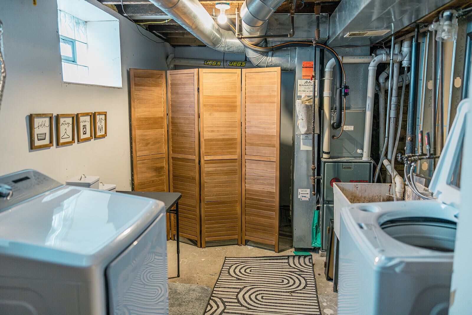 This functional laundry area includes a washer, dryer, and utility sink, with a wooden folding screen for privacy. Exposed pipes add an industrial touch, while framed wall art enhances the space. A practical, well-organized spot for guests’ convenience.