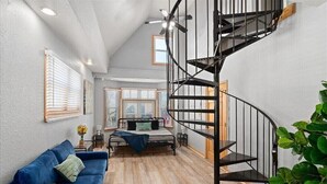A bedroom with a bed, a blue sofa, a wooden table, a black spiral staircase, and a large window with blinds. - 𖤓GrandVictorian 5000sqft Sale Sleeps23 ArcadeGameFloor BBQ𖤓 (Milwaukee)