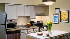 Una cocina moderna con armarios blancos, electrodomésticos de acero inoxidable, encimera de granito y una zona de comedor con una mesa blanca y sillas a rayas.