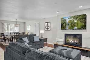 A modern living room with a fireplace, a dining area with a table and chairs, and a large window with blinds.