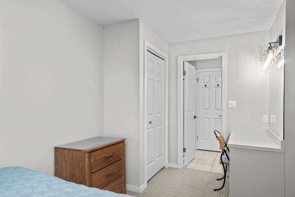 Un dormitorio con cama, cómoda de madera, espejo y una puerta que da a otra habitación.