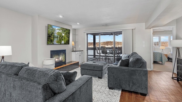 A modern living room with a fireplace, grey sofas, a dining area with outdoor seating, and a large window overlooking a natural landscape.