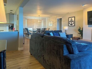 Een moderne woonkamer met een donkergrijze bank, houten vloeren, een eethoek met een tafel en stoelen, en een keuken met een koffieapparaat.