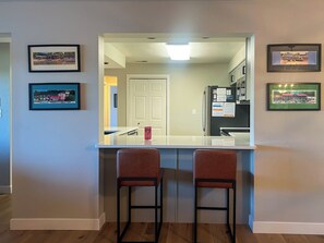 Een keuken met een wit aanrecht, twee barkrukken en lijstjes met foto's aan de muur.