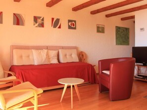 Un salon chaleureux avec un canapé rouge, une petite table ronde et un fauteuil rouge. Des tableaux abstraits ornent le mur, et un téléviseur est posé sur un meuble.
