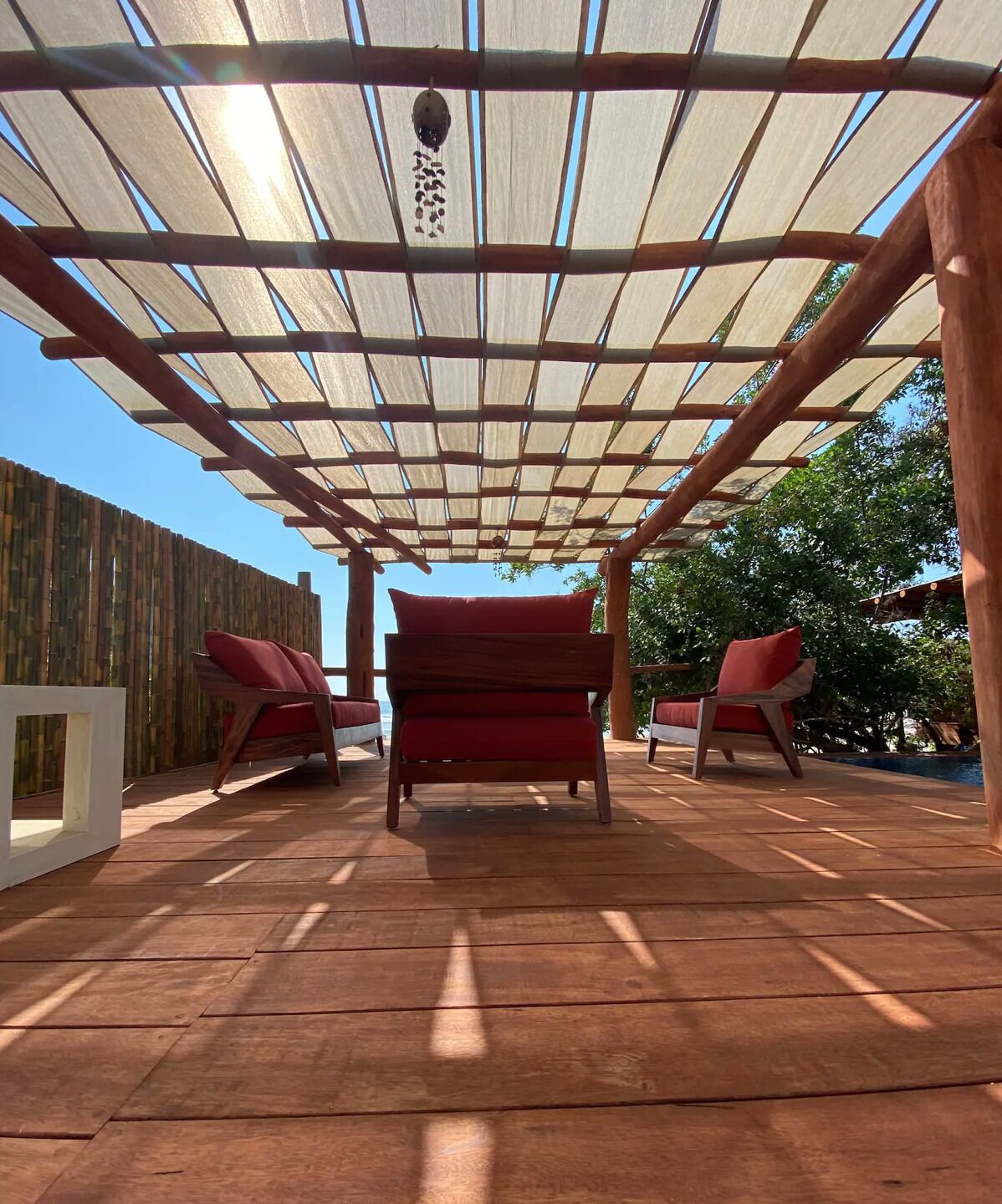 A wooden deck with a pergola overhead, two red cushioned chairs, and a wooden table.