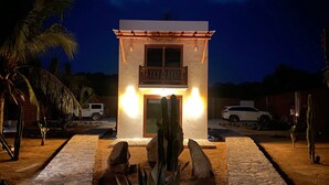 A small, two-story building with a sign reading "Casa de la Vida." It features a wooden balcony, a stone pathway leading to the entrance, and is surrounded by cacti and palm trees. - Amazing private beachfront Village (Troncones)