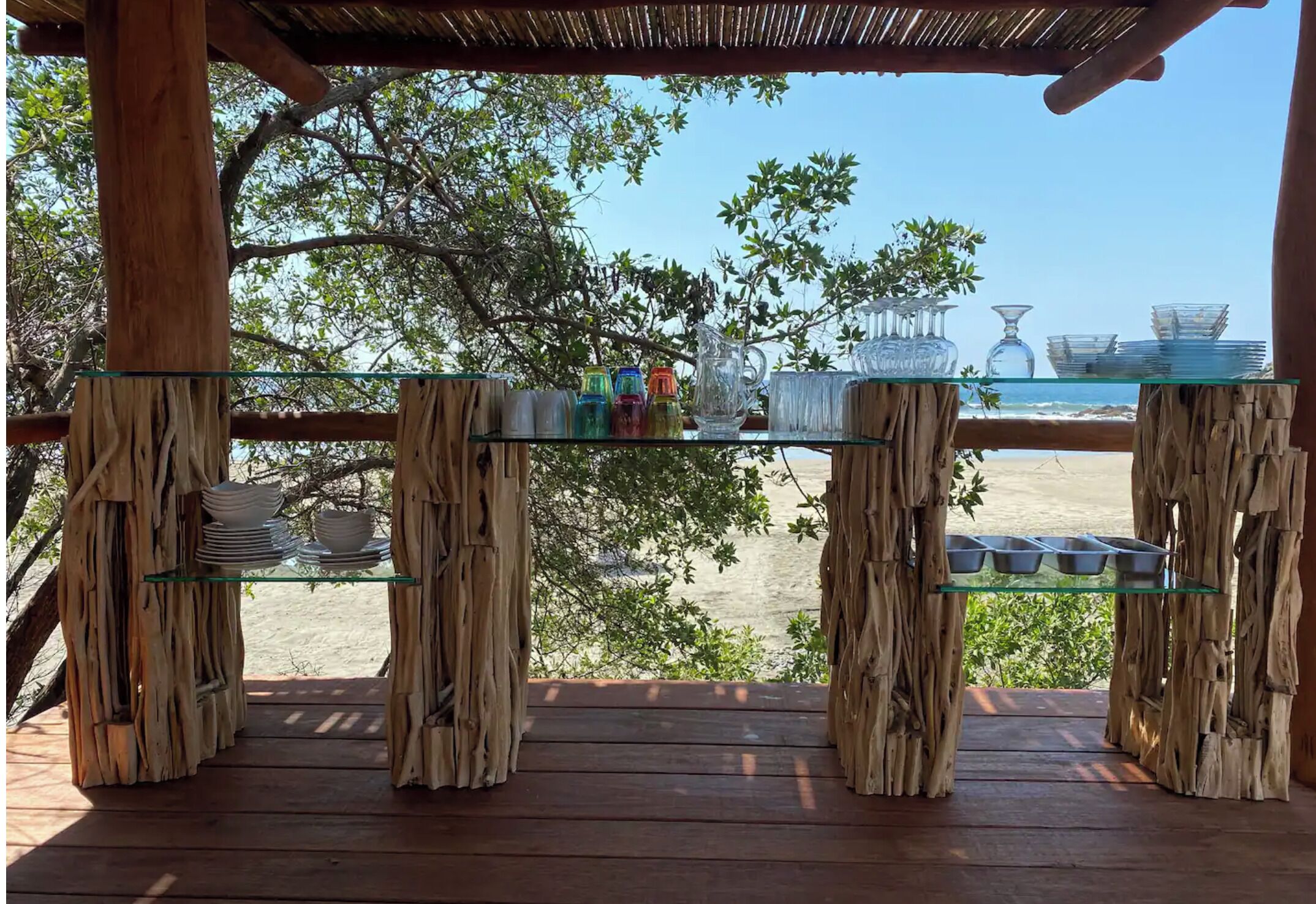 A rustic outdoor seating area with a wooden deck and glass tables, overlooking a beach.
